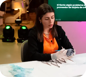 Dra. Rayane en camisa naranja y chaqueta negra con guantes blancos está examinando un objeto gris con herramientas dispuestas sobre una mesa blanca.