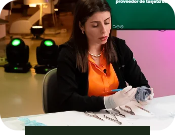 Dra. Rayane en camisa naranja y chaqueta negra con guantes blancos está examinando un objeto gris con herramientas dispuestas sobre una mesa blanca.